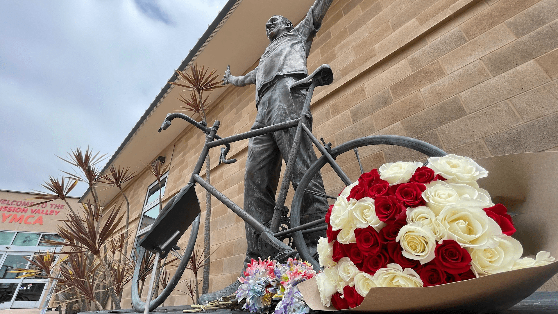 Flowers left outside of the Mission Valley YMCA on May 27, 2024. There is a statue of Bill Walton with his bike and a plaque that reads, “The Y saved my life.”