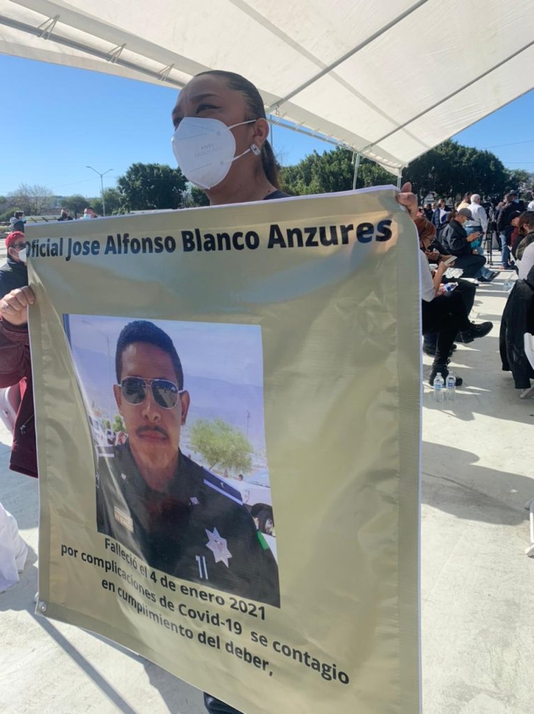 mujer con cubrebocas sostiene letrero con foto de oficial que murio de covid