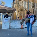 Una madre carga a su hijo de 14 años frente a la frontera entre Tijuana y San Diego con alambrado de púas. Dos agentes con cubrebocas esperan a un lado.