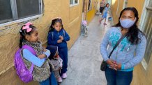 Mujer con cubrebocas en un albergue en Tijuana de color amarillo con dos niñas bien peinadas con moños rosas y un niño pequeño viendo a una de ellas. Una niña y dos señoras están al fondo mirando a la cámara