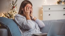 A sad woman sitting on a couch on a laptop with a Christmas tree in the background.