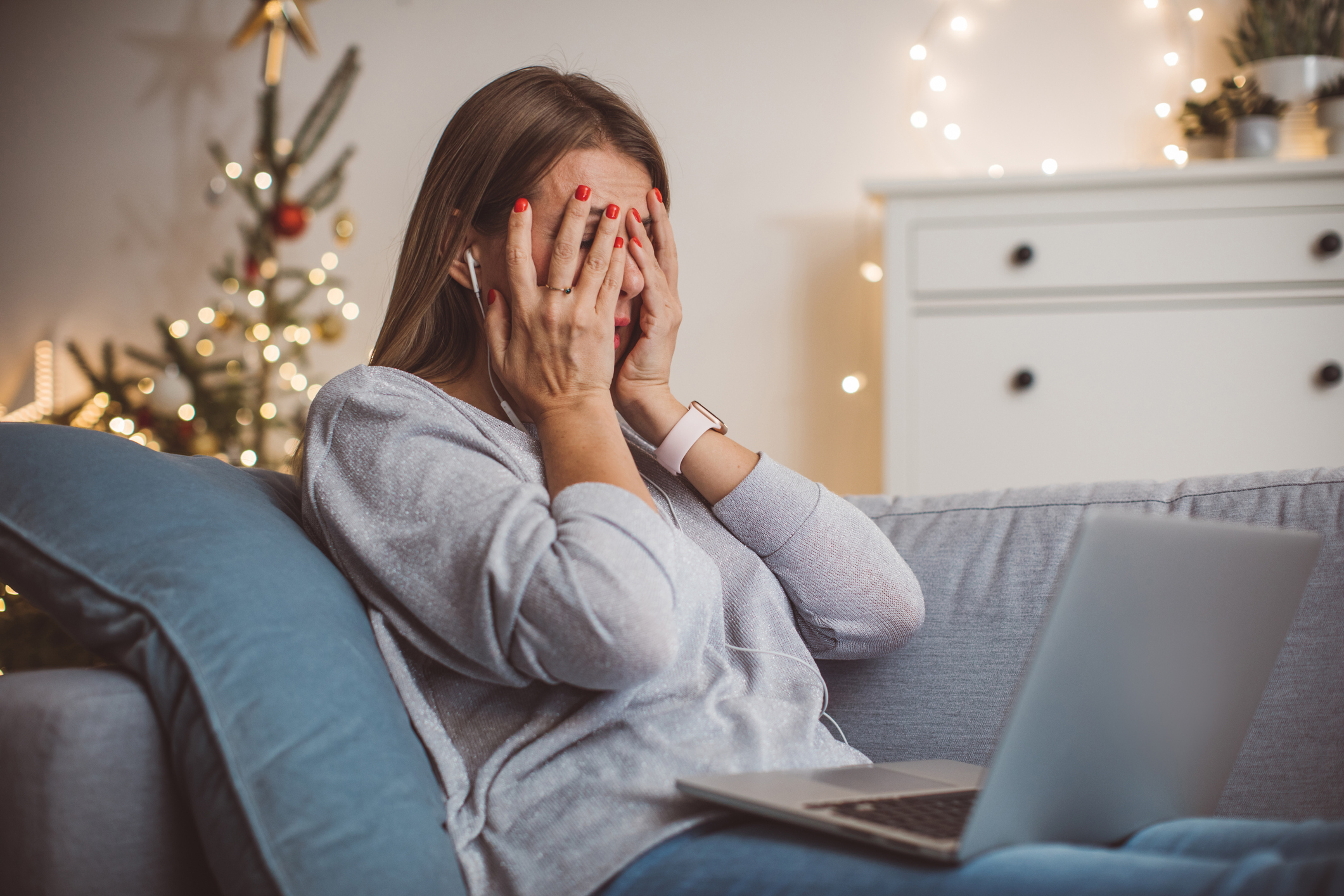 A sad woman sitting on a couch on a laptop with a Christmas tree in the background.