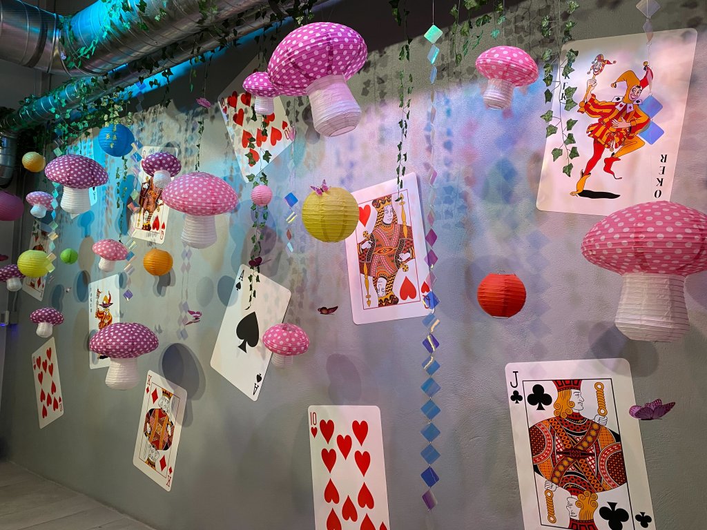 Paper mushrooms and balls dangle in front of a background of decorative cards on a wall at The Alice - An Immersive Cocktail Experience.