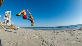 A woman doing a flip into the air off of a cement wall onto the sand of the beach while working out in the spring in the city of San Diego, California