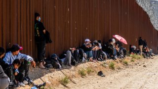 MIgrantes sentados al lado del muro fronterizo.