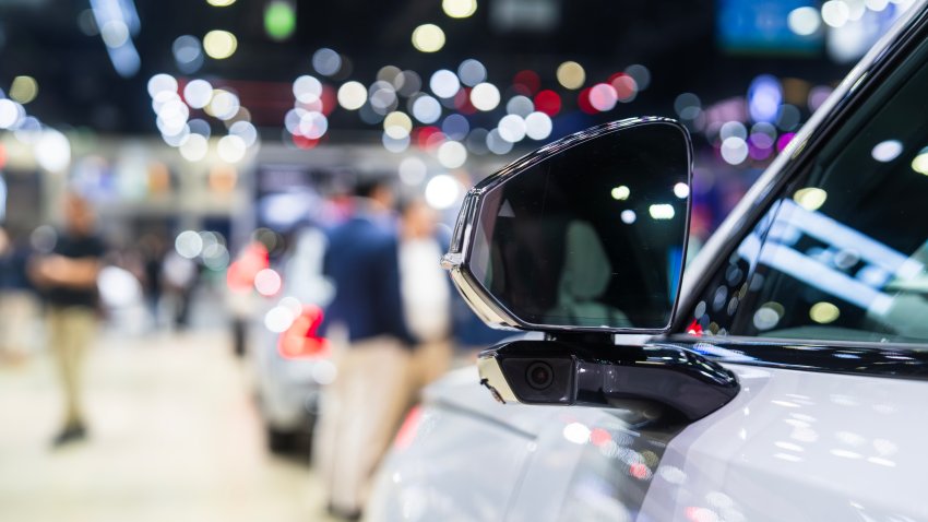 A detailed close-up of a car mirror at a bustling auto show, highlighting the modern design. The blurred background features colorful bokeh lights, adding a vibrant and lively atmosphere.